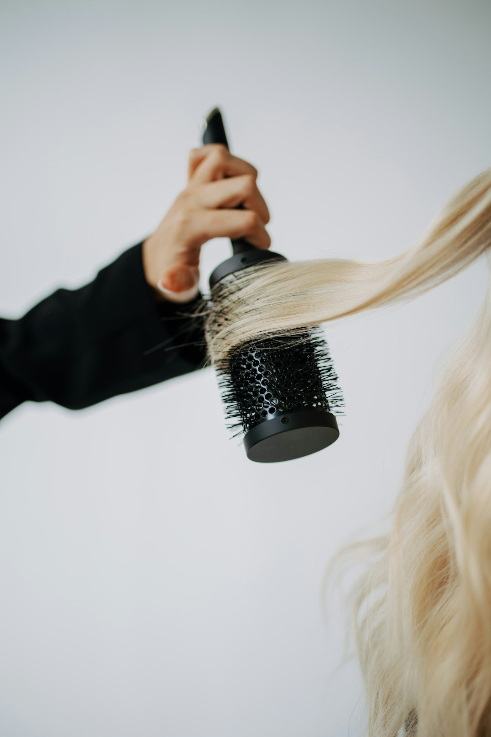 Mão segurando escova térmica ao estilizar cabelo loiro comprido